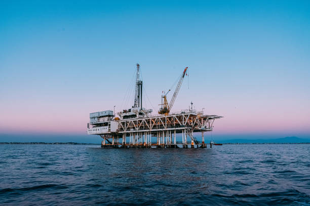 A striking image of a perfectly centered offshore oil rig at sunset off the coast of Huntington Beach, California. The pink & orange tones of the setting sun highlight the industrial machinery and equipment used in the drilling and extraction of fossil fuels, including crude oil and natural gas. 

The scene captures the intersection of the energy industry and the beauty of the Pacific Ocean. The image speaks to issues of fuel and power generation, energy crises, and environmental concerns surrounding the oil and gas industry.